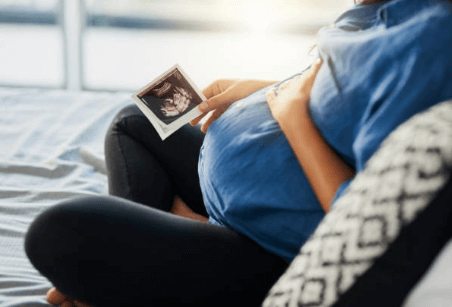 Woman sitting on a cozy bed with her hand on her pregnant belly and holding an ultrasound image in the other hand.
